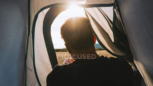 Back view of crop unrecognizable male traveler in casual clothes lying in camping tent and admiring picturesque mountain landscape on sunny day — Stock Photo