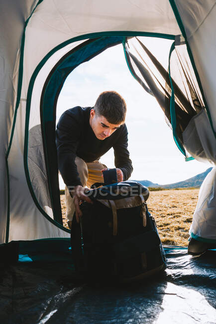 Jovem elegante macho campista em roupa casual colocando mochila dentro de tenda durante caminhadas viagem no vale montanhoso em luz solar — Fotografia de Stock