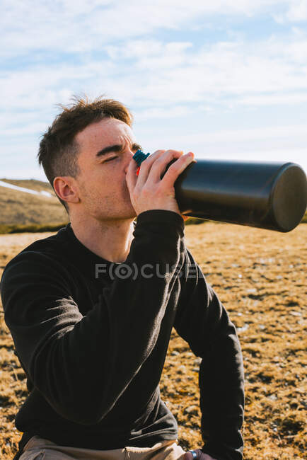 Side view of young stylish male traveler in casual outfit sitting on haunches near backpack and drinking hot beverage from thermos during hiking trip in mountainous terrain — Stock Photo