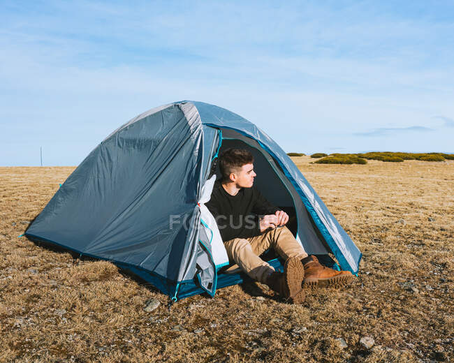 Corpo pieno di moda giovane viaggiatore maschile in abito elegante seduto in tenda da campeggio durante il riposo dopo il trekking — Foto stock