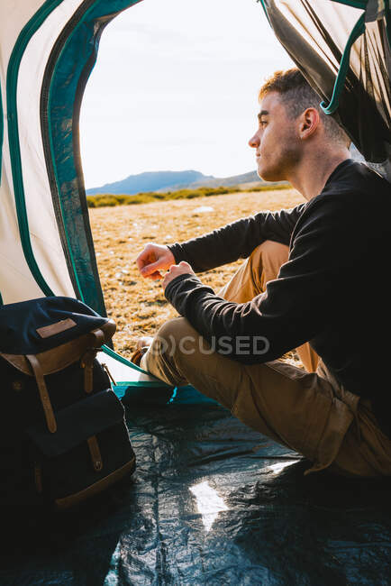 Side view of young dreamy male traveler in casual outfit resting in camping tent and admiring picturesque mountainous terrain on sunny day — Stock Photo