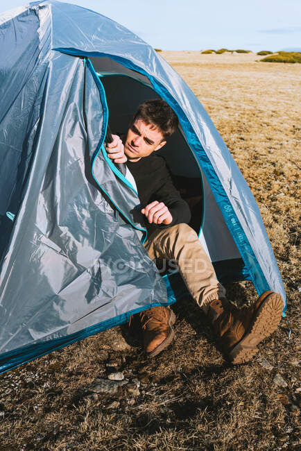 Ganzkörper trendiger junger männlicher Reisender in stylischem Outfit sitzt nach dem Trekking im Zeltlager und ruht sich aus — Stockfoto