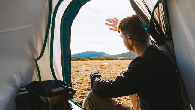 Back view of young dreamy male traveler in casual outfit resting in camping tent and admiring picturesque mountainous terrain on sunny day — Stock Photo