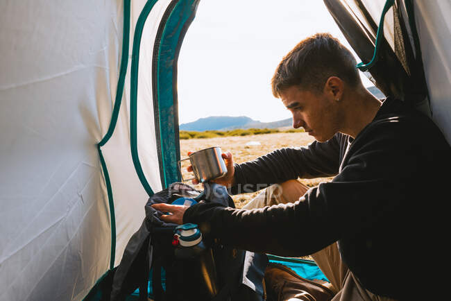 Side view of calm young male camper inc casual clothes enjoying hot drink while resting in tent after hiking in mountains on sunny day — Stock Photo