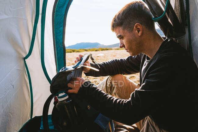 Side view of young dreamy male traveler in casual outfit resting in camping tent and checking his backpack on sunny day — Stock Photo