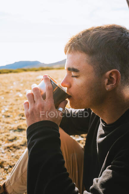 Fiducioso giovane escursionista di tendenza maschile in abito caldo bere tazza di bevanda calda, mentre ricreando in tenda campeggio nella giornata di sole — Foto stock