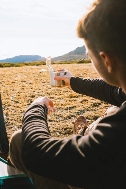 Vista posteriore di fiducioso giovane escursionista di tendenza maschile in abito caldo bere tazza di bevanda calda, mentre ricreando in tenda campeggio nella giornata di sole — Foto stock