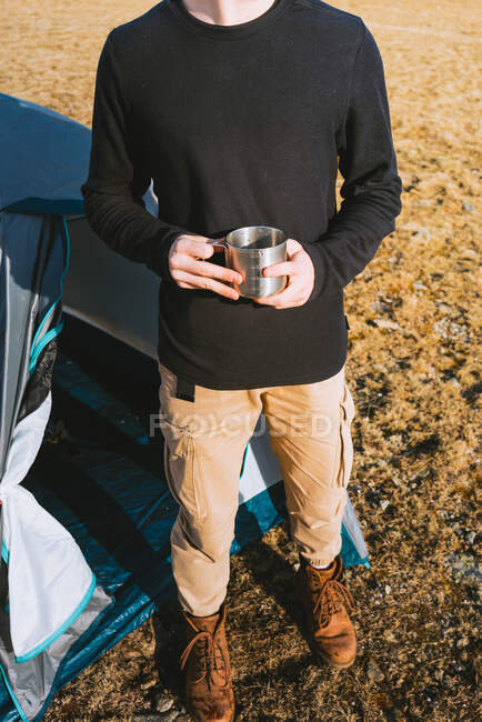 Анонимный молодой модный турист в теплой одежде, пьющий кружку горячего напитка во время отдыха в палатке в солнечный день — стоковое фото