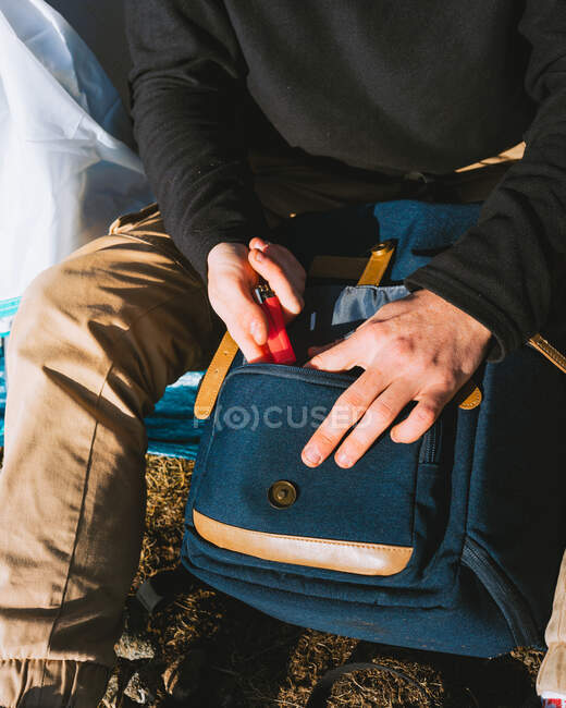 Desde arriba de la cosecha caminante masculino anónimo en traje casual poner encendedor en el bolsillo de la mochila mientras está sentado en la tienda de campaña durante el viaje en el día soleado - foto de stock