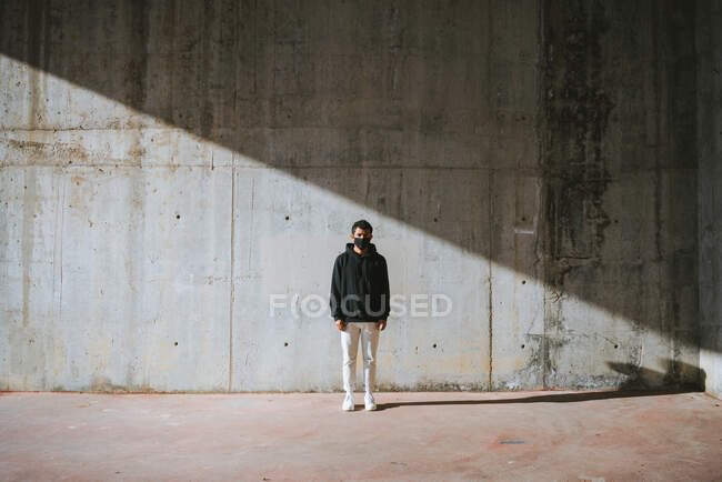 Unrecognizable young male in hoodie and mask standing on street near concrete wall — Stock Photo