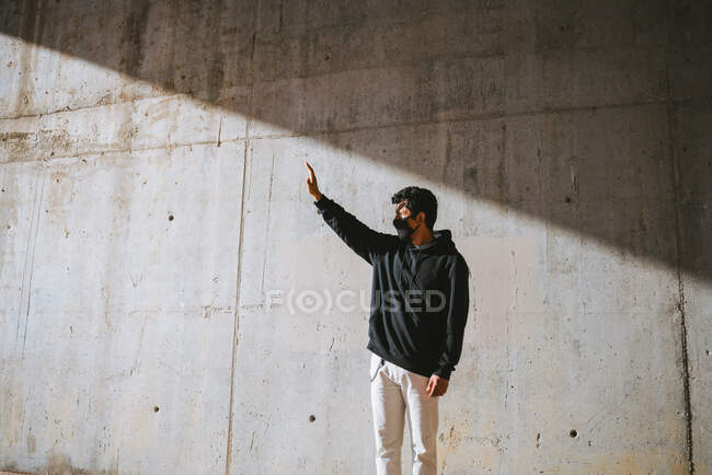 Неузнаваемый молодой мужчина в толстовке и маске стоит на улице возле бетонной стены и прикрывает лицо рукой от яркого солнечного света — стоковое фото