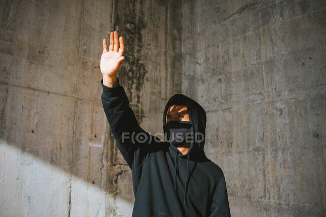 Hombre joven irreconocible con capucha y máscara de pie en la calle cerca de la pared de hormigón y cubriendo la cara con la mano de la luz del sol brillante - foto de stock