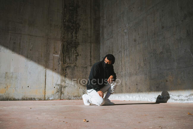 Неузнаваемый молодой мужчина в толстовке и маске сидит на улице возле бетонной стены — стоковое фото