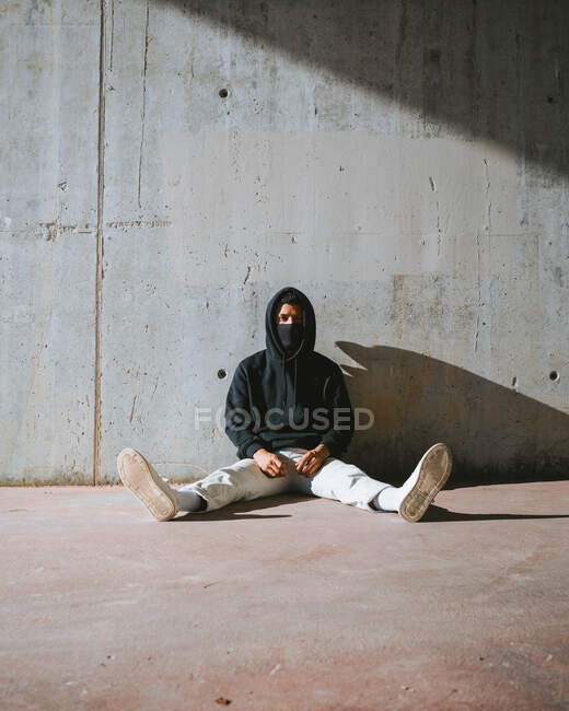 Jeune homme méconnaissable en sweat à capuche et masque assis sur la rue près d'un mur de béton — Photo de stock
