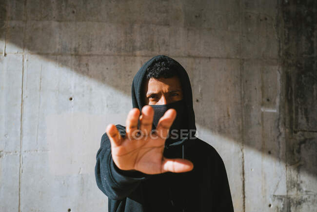 Young male in hoodie and mask standing on street near concrete wall and hand up — Stock Photo