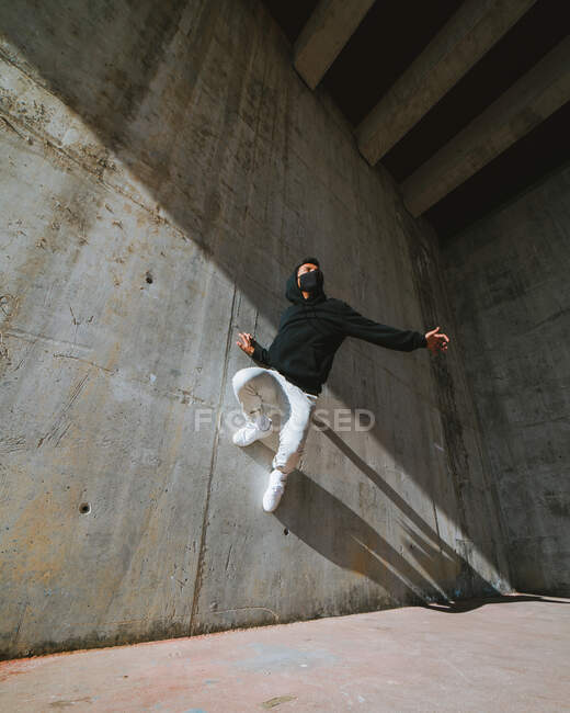 Von unten ein unkenntlich gemachter junger Mann in modischem Outfit und Maske, der bei einem Tanz in einem verlassenen Gebäude an einem sonnigen Tag einen Trick an einer Betonwand vorführt — Stockfoto
