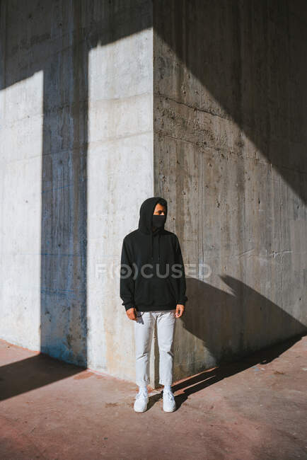 Trendiger Typ mit Gesichtsmaske, der an sonnigen Tagen auf der Straße in der Nähe einer schäbigen Mauer steht und wegschaut — Stockfoto