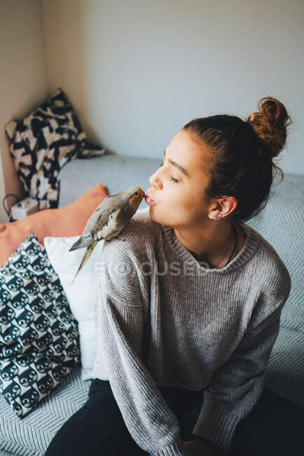 Joven propietaria en ropa casual preparándose para besar adorable pájaro cacatúa sentado en el hombro en la sala de estar - foto de stock