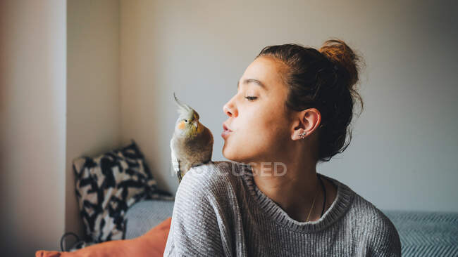Young female owner in casual clothes preparing to kiss adorable cockatiel bird sitting on shoulder in living room — Stock Photo