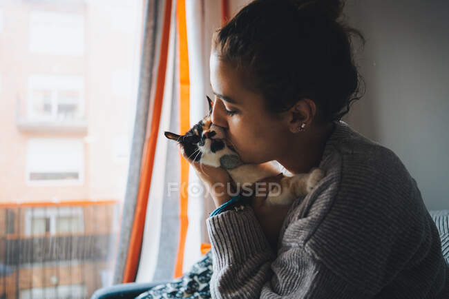 Side view of young female owner in casual outfit hugging and kissing cute fluffy calico cat with closed eyes while resting on sofa in living room — Stock Photo