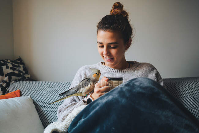 Felice signorina in maglione caldo sorridente e bere caffè caldo mentre si rilassa sul divano con adorabile uccello cockatiel a portata di mano — Foto stock