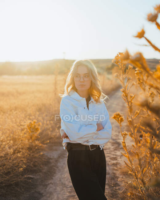 Vue latérale de la jeune voyageuse confiante en vêtements décontractés et lunettes debout dans la prairie et regardant loin tout en se relaxant à la campagne au coucher du soleil — Photo de stock