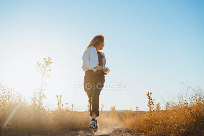 Vista frontal de jovem turista na moda com cabelo loiro em roupas casuais e óculos correndo na estrada rural em meio a plantas secas no campo — Fotografia de Stock