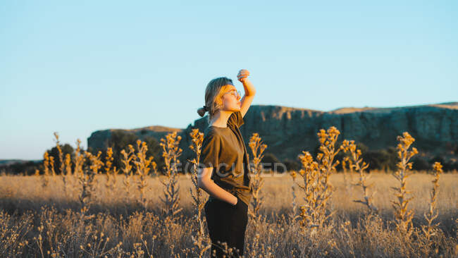 Side view of confident young female traveler in casual clothes and eyeglasses standing in meadow and looking away while relaxing in countryside at sunset — Stock Photo