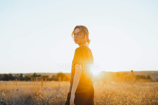 Side view of confident young female traveler in casual clothes and eyeglasses standing in meadow and looking t camera while relaxing in countryside at sunset — Stock Photo