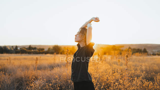 Вид сбоку молодой девушки в повседневной одежде, наслаждающейся летним днем, стоя на сухом поле с поднятыми руками на закате в сельской местности — стоковое фото