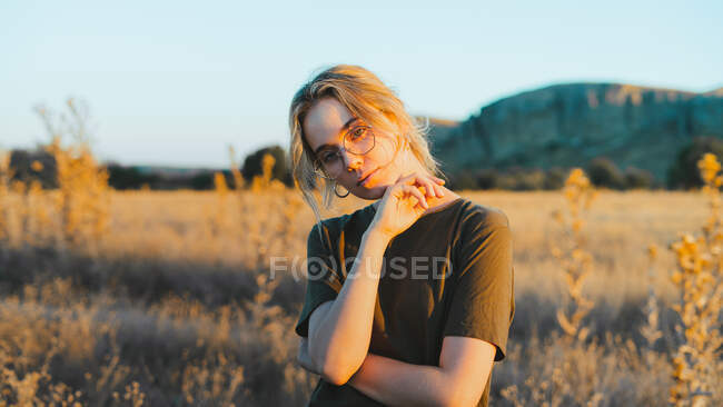 Front view of confident young female traveler in casual clothes and eyeglasses standing in meadow and looking away while relaxing in countryside at sunset — Stock Photo