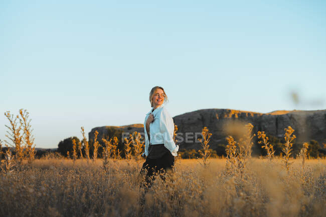 Seitenansicht einer jungen Dame mit blonden Haaren in stilvoller Kleidung, die inmitten von Gras auf einem ländlichen Feld in der Nähe von Hügeln gegen wolkenlosen blauen Himmel bei Sonnenuntergang spaziert — Stockfoto