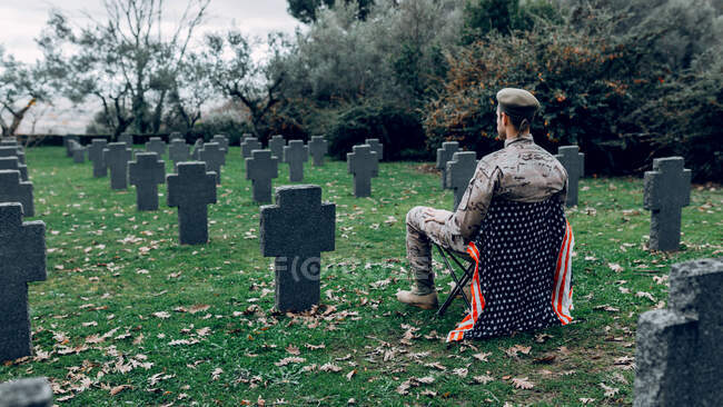 Back view full body of soldier in uniform sitting on chair with American flag while mourning death of warriors at graveyard — Stock Photo