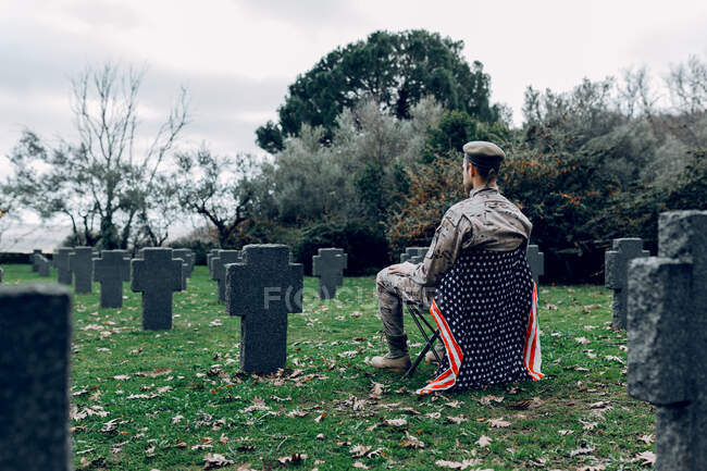 Vista posterior cuerpo completo de soldado en uniforme sentado en silla con bandera americana mientras lloraba la muerte de los guerreros en el cementerio - foto de stock