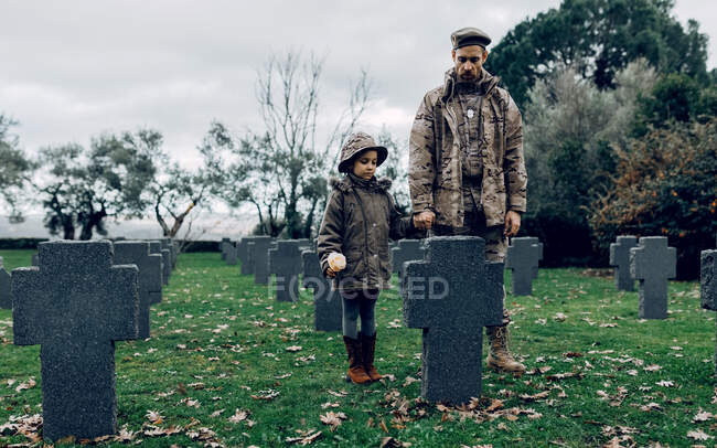 Corpo inteiro de guerreiro masculino de mãos dadas com a menina sentindo tristeza pelo herói falecido no cemitério — Fotografia de Stock
