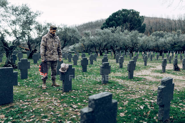 Pieno corpo di soldato in uniforme in piedi con bandiera americana mentre piange la morte dei guerrieri al cimitero — Foto stock