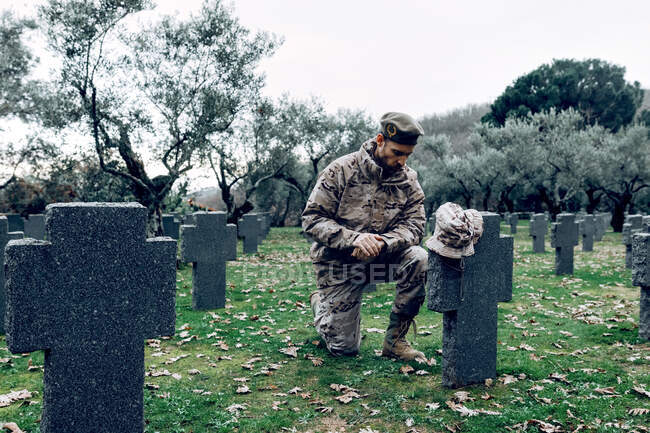 Soldado entristecido de cuerpo entero disfrazado de camuflaje arrodillado frente a la tumba en el cementerio militar a principios de otoño - foto de stock