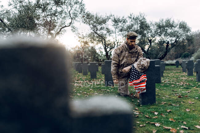 Soldato pieno corpo doloroso in abito camuffamento inginocchiato davanti alla tomba nel cimitero militare all'inizio della giornata autunnale — Foto stock