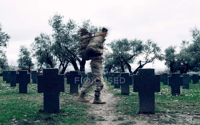 Vista lateral soldado anónimo en camuflaje llevando mochila guerrera caminando en amplio cementerio militar - foto de stock