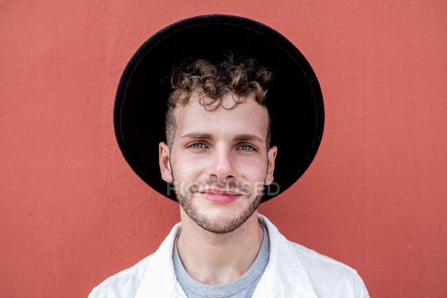 Joyeux jeune homme millénariste en vêtements élégants souriant tout en se reposant sur la rue contre le mur rouge — Photo de stock