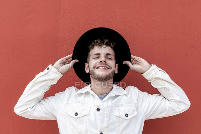 Веселый молодой мужчина тысячелетия в стильной одежде регулируя шляпу и улыбаясь с закрытыми глазами во время отдыха на улице у красной стены — стоковое фото
