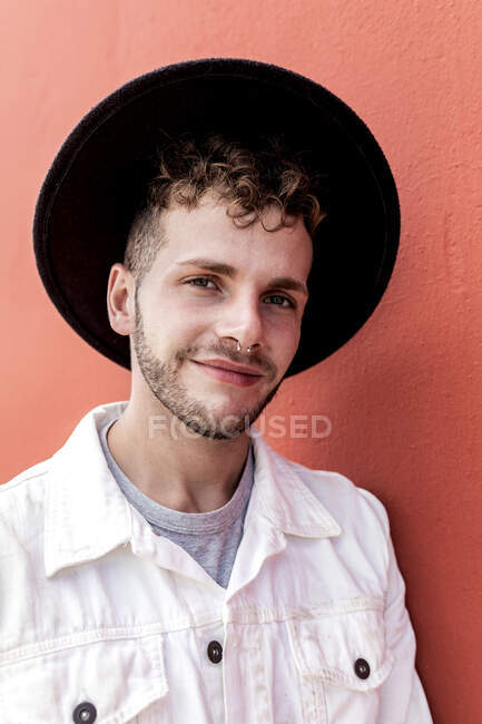 Cheerful young male millennial in stylish clothes smiling while resting on street against red wall — Stock Photo