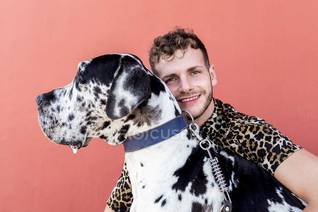 Jeune homme non rasé en vêtements décontractés et adorable Arlequin obéissant Grand Dane chien étreignant l'autre sur fond rouge — Photo de stock