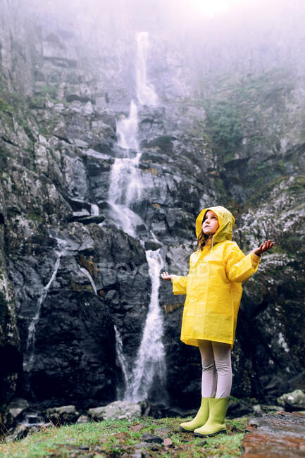 Девушка-подросток в ярком дождевике с поднятыми руками смотрит на каскад с быстрым потоком воды на горе — стоковое фото