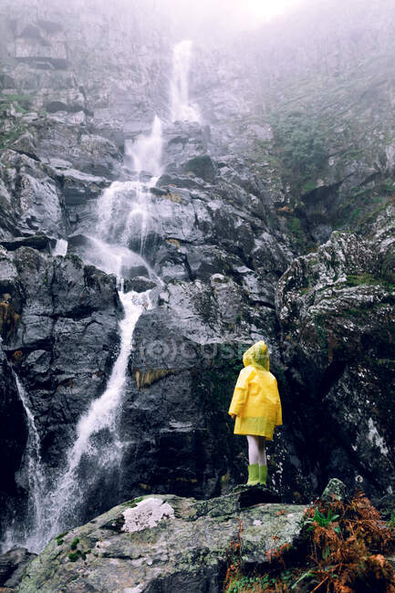Vue arrière d'une adolescente méconnaissable en imperméable brillant debout contre la cascade avec écoulement d'eau rapide sur le mont — Photo de stock