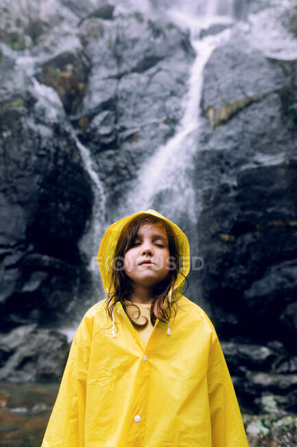 Female teenager in bright raincoat standing with eyes closed against cascade with fast water flow on mount — Stock Photo