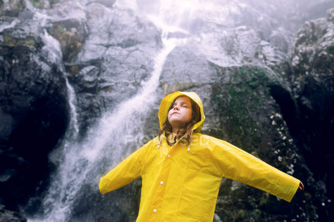 Женщина-подросток в ярком дождевике с закрытыми глазами и поднятыми руками, стоящая против каскада с быстрым потоком воды на горе — стоковое фото