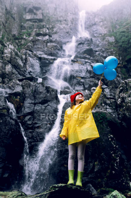 Cheerful teen in raincoat with balloons looking up against high mountain with foamy water cascade during trip — Stock Photo