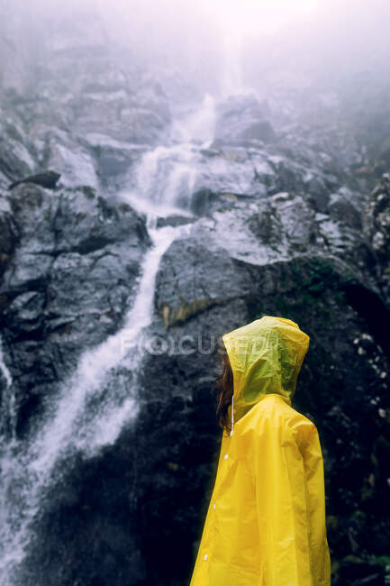 Вид сбоку на неузнаваемую девочку-подростка в ярком дождевике, стоящую напротив каскада с быстрым потоком воды на горе — стоковое фото