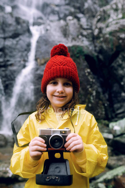 Delícia adolescente em capa de chuva tirar foto na câmera contra montagem áspera com cascata espumosa à luz do dia — Fotografia de Stock